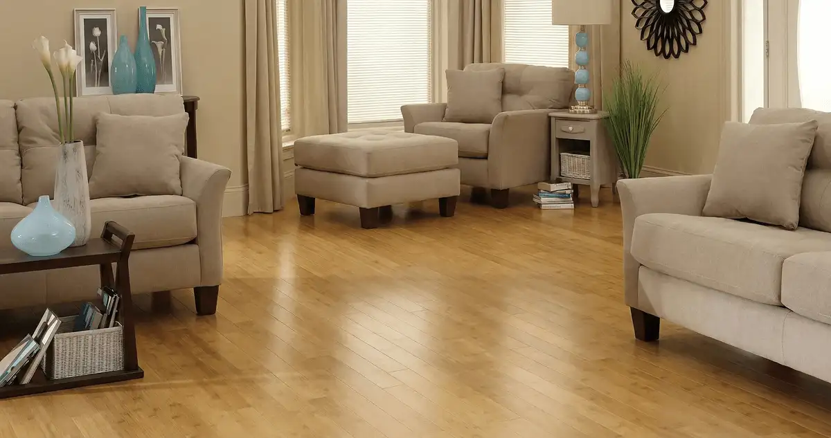 A living room with professionally installed bamboo flooring, beige sofas, a matching ottoman, a side table with a blue lamp, vases, framed pictures, and large windows with blinds and curtains in Orange, CA